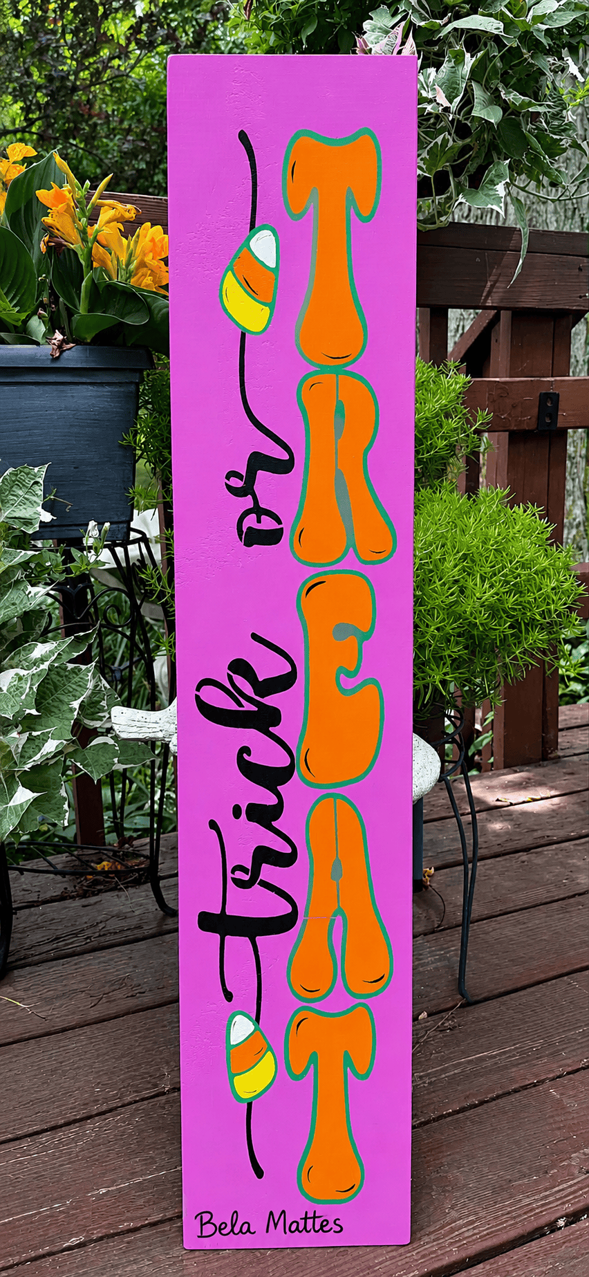 A tall rectangular Halloween sign with a bright pink background, featuring the words 'Trick or Treat' in black script and large orange letters outlined in green that spell 'TREAT'. Small candy graphics appear near the top and bottom, and the name 'Bela Matte' is printed at the bottom.
