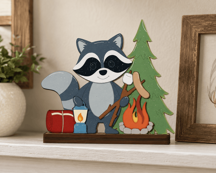 A decorative wooden cutout of a raccoon camping by a tree, shown on a shelf in a styled indoor setting with framed art and a plant.